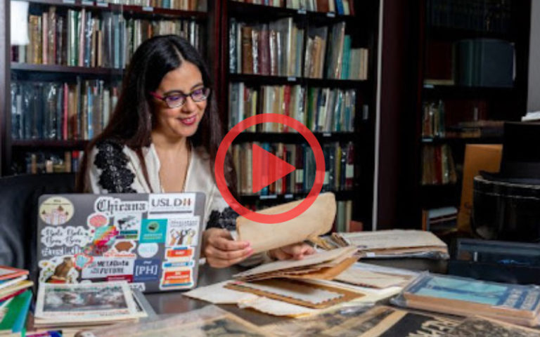 Lorena Gathereau sits reading at table filled with newspapers and laptop in library.