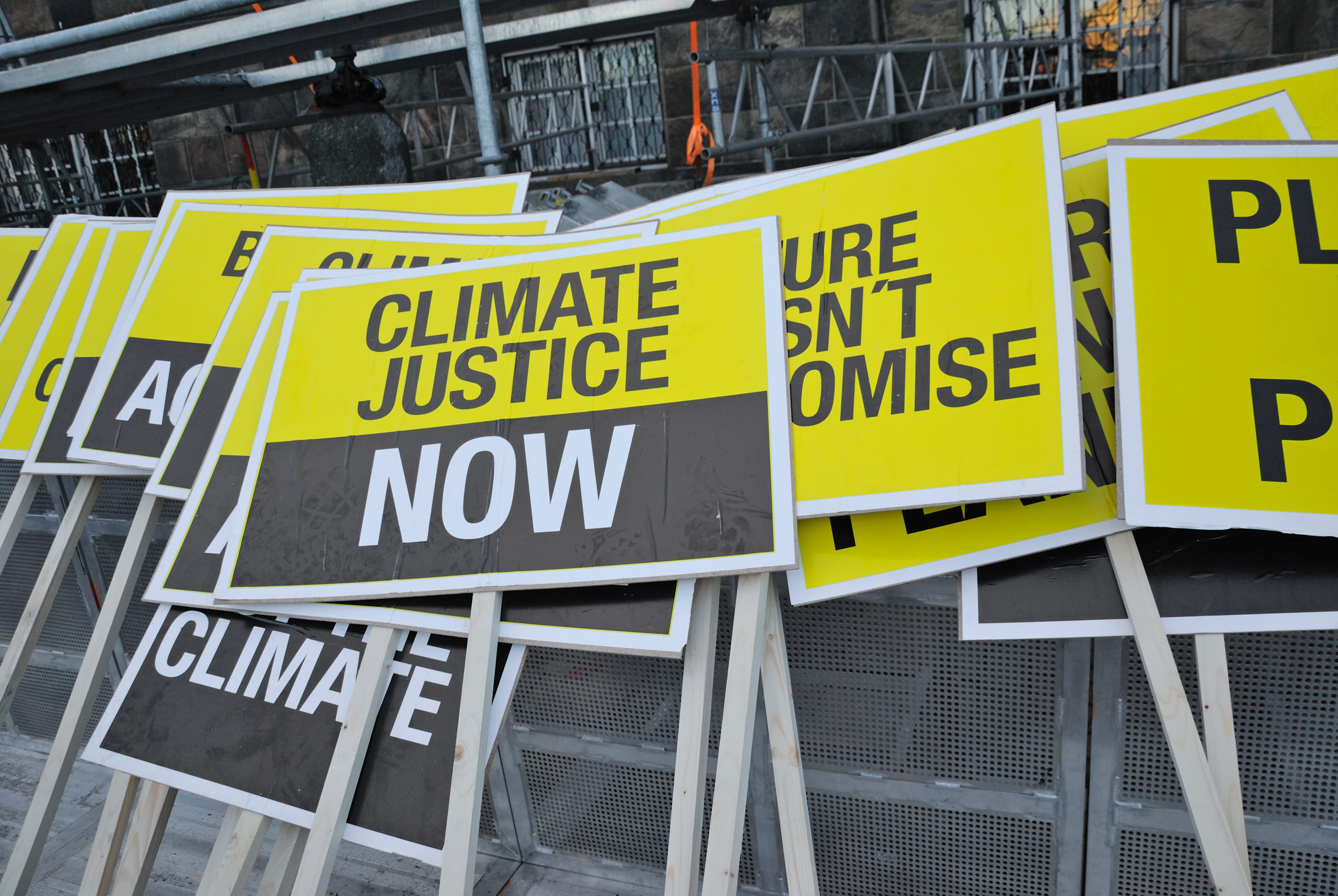 Color photo of pile of yellow and black protest signs reading "Climate Justice NOW."