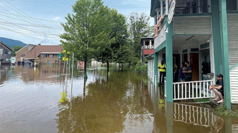 Color photo of flooded small town square. Side view of home with double decker porch filled with stranded flood victims waiting for water to recede.