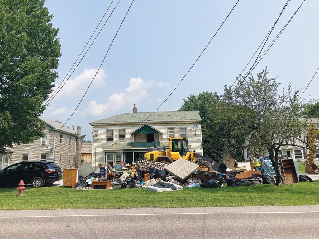 Color photo of across the street view of three homes with flood soaked content and debris in large pile on yard.