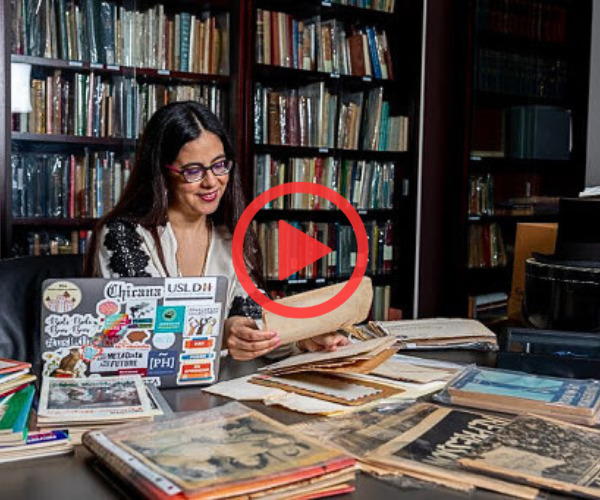 Lorena Gathereau sits reading at table filled with newspapers and laptop in library.