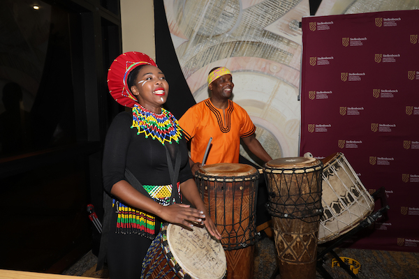 Color photo of two people standing and playing traditional South African drums.