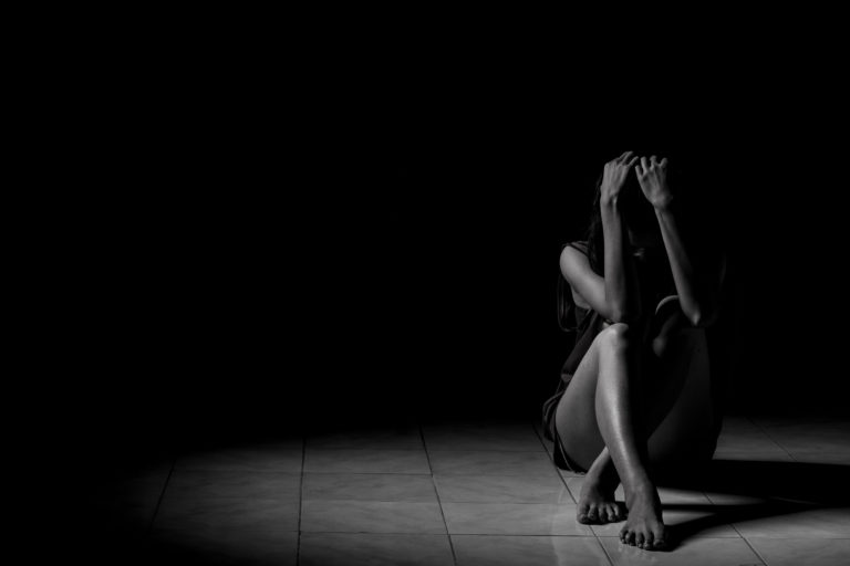 Black and white photo of a depressed woman sitting on floor alone in dark room with knees supporting arms and hands that cover her face.