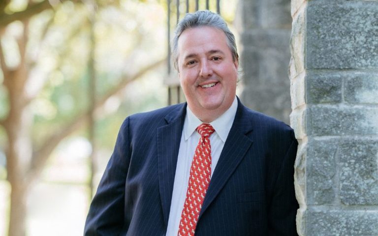 Color photo portrait of Joey King standing outside, leaning on stone wall, smiling at camera while wearing dark suit with red tie.