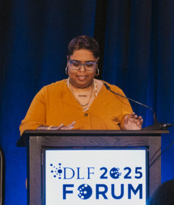 The photo shows a person in an orange dress speaking at a podium that reads "DLF 2025 Forum."