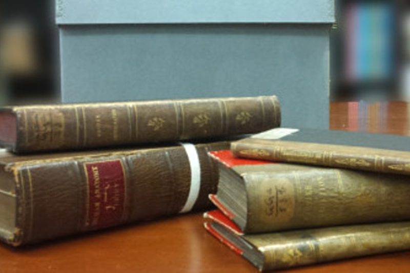 A set of old books of varying sizes on a table in the foreground. Archival boxes in the background.